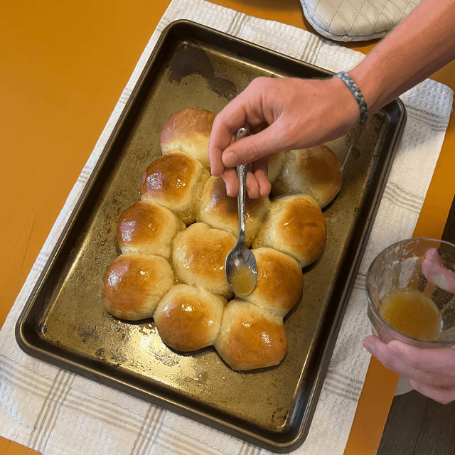 Pouring honey over baked breadrolls in a pan with a spoon.